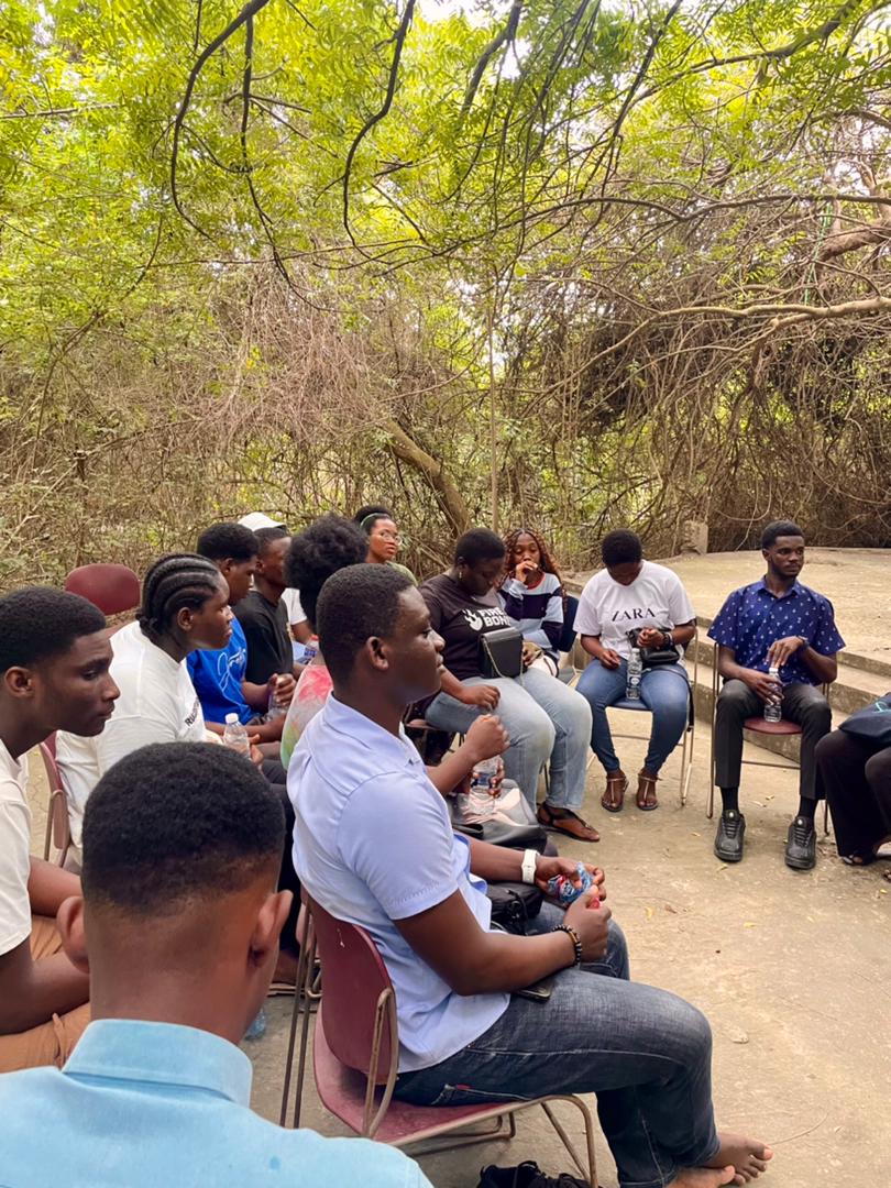 A Kings & Priests duo laugh together, one seated backward on a chair wearing a red vest with a yellow sickle-and-hammer symbol and socks reading “Vegan vibes.”