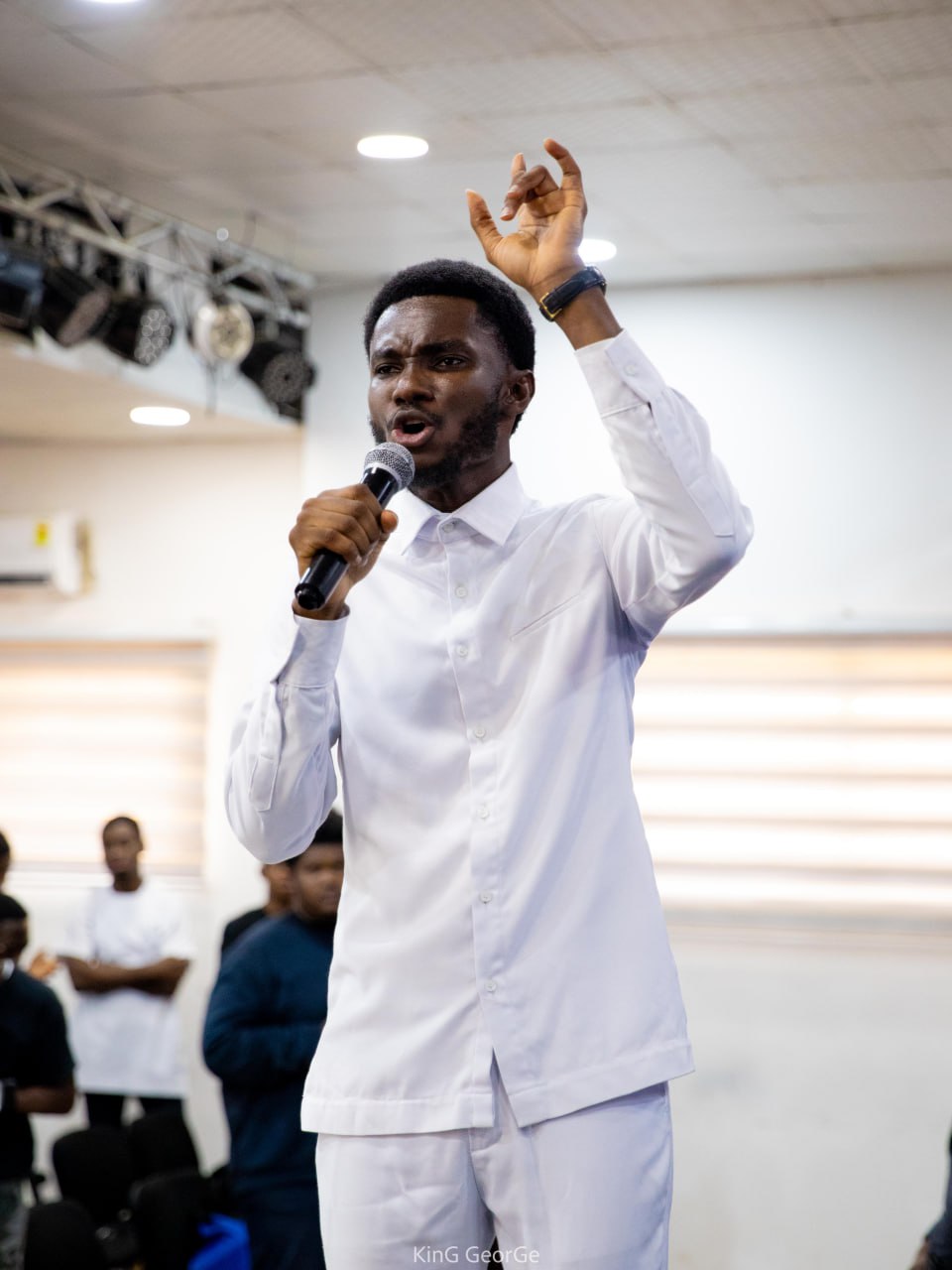 A Kings & Priests worshipper stands peacefully in a floral hoodie against a pink background, eyes closed in prayerful rest.